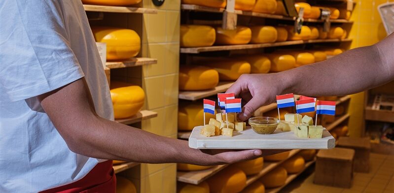 Een medewerker van de Gouda Cheese Experience overhandigt een plankje met kaasblokjes en mosterdsaus, versierd met Nederlandse vlaggetjes, aan een bezoeker. Op de achtergrond zijn rekken vol Goudse kazen te zien.