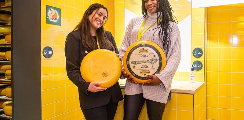Twee bezoekers poseren lachend met grote Goudse kaaswielen in een gele interactieve ruimte bij de Gouda Cheese Experience.