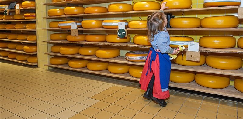 Kind in kaasoutfit reikt naar een grote kaaswiel in een opslagruimte vol Goudse kazen bij de Gouda Cheese Experience
