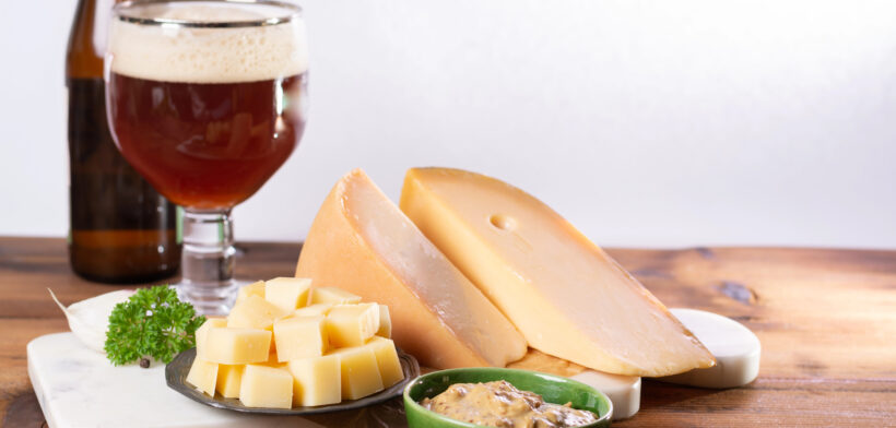 Glas ambachtelijk bier met blokjes Goudse kaas, kaaswiel en dip op een houten tafel.